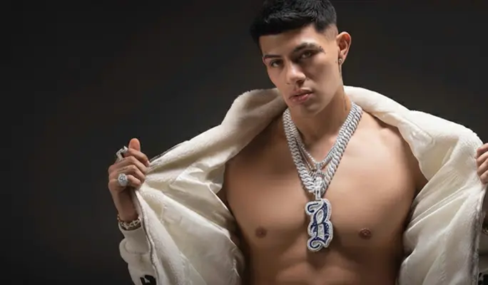 El Bogueto, shirtless and wearing heavy silver chains, looking directly at the camera while wearing a white fluffy jacket over his shoulders.