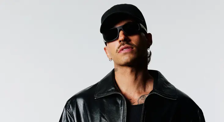 Reggaeton artist Feid wearing a black baseball cap, black sunglasses, and a black leather jacket, promoting his track Princesa.