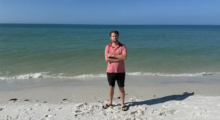 Producer J.M. Dee standing on a white sand beach with the ocean in the background.