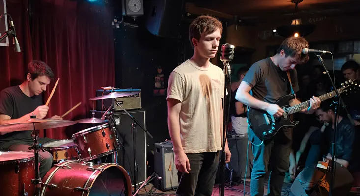 A three-piece alternative rock band performing on a small, intimate stage with a drummer, lead singer, and guitarist.