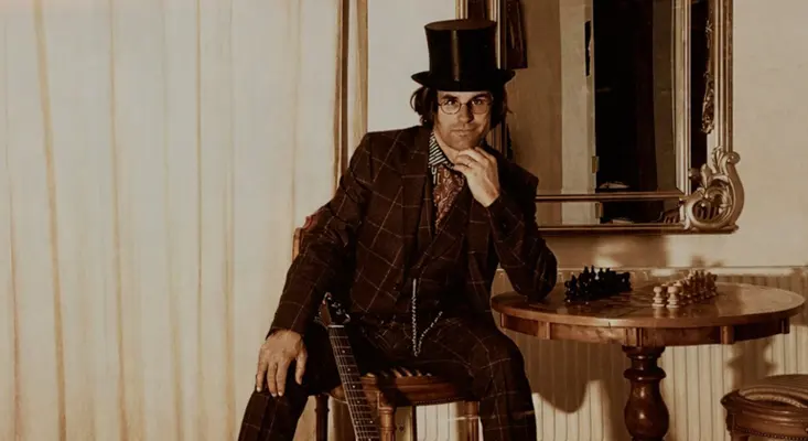 The ‘Dandyworld’ Album: An Unfiltered Interview With Dandy John Musical artist Dandy John wearing a top hat and suit, sitting next to a chessboard with a guitar, representing his Dandyworld persona.