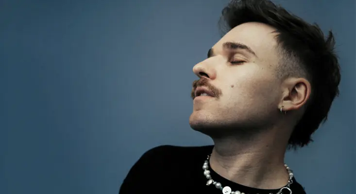 Ely Oaks looking upward with eyes closed against a blue background, wearing a black top and pearl necklace.