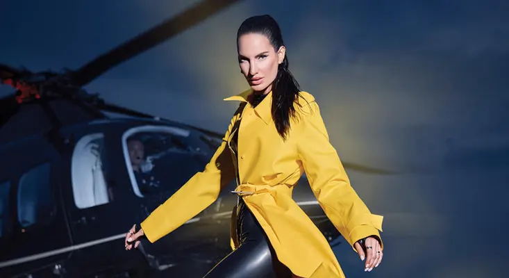 Lilly Palmer standing in front of a luxury helicopter wearing a yellow trench coat in the Bigger Than Techno music video.