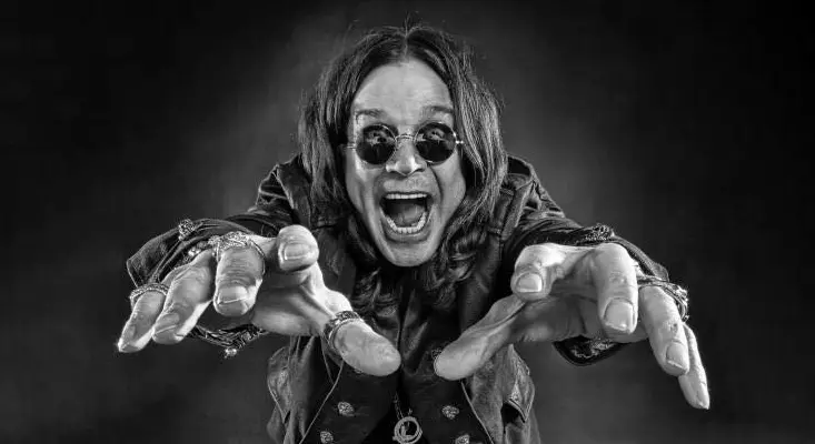 Ozzy Osbourne in a dramatic black and white portrait with hands outstretched, round sunglasses and long hair, the original artist behind Crazy Train.