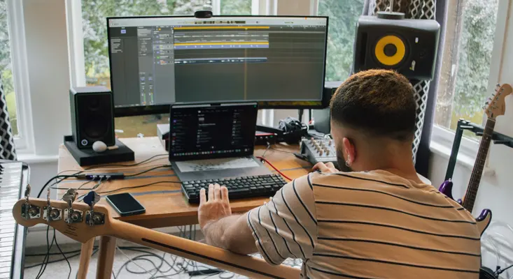 A music producer working on a DAW in a modern home studio setup with monitors, guitar, and mixer.