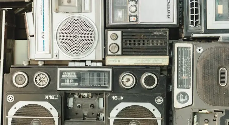 From "Madchester" to modern subgenres, music is a living ecosystem. These vintage players represent the tangible roots of an ever-evolving cultural history. A collection of vintage boomboxes and cassette recorders stacked together, symbolizing the evolution of music culture and physical media.