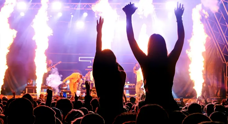 A large, diverse crowd with hands raised at a live concert with pyrotechnics, illustrating how dual-genre artists reach multiple fan bases.