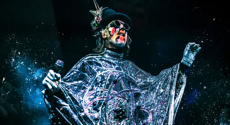 Arthur Brown performing live in a shimmering silver robe and top hat with theatrical face paint, demonstrating his pioneering influence on Shock Rock.