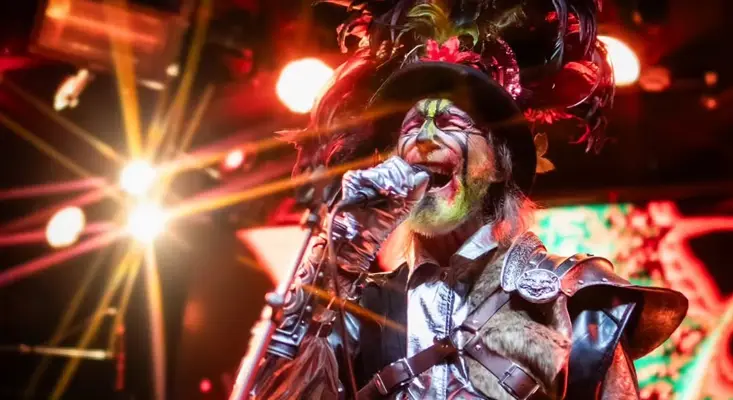Arthur Brown singing with intense expression, wearing a flamboyant feathered hat and metallic stage gear, representing his experimental and surreal Rock legacy.