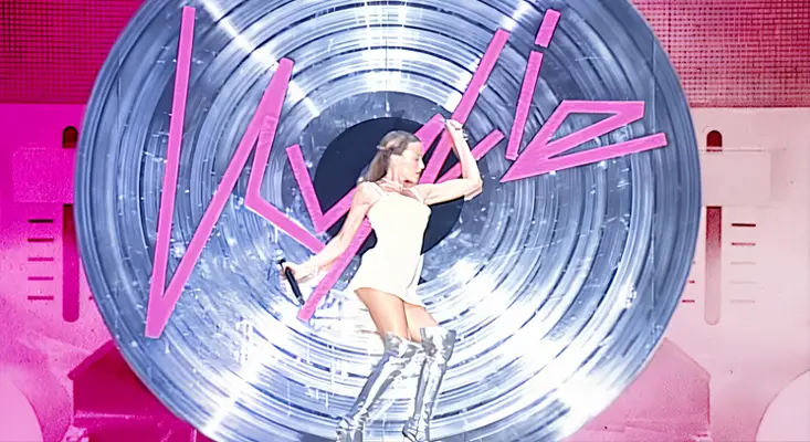 Kylie Minogue performing on stage in a white outfit and silver boots in front of a giant spinning disc with her name in pink lights featured in the Kylie Minogue Netflix documentary.
