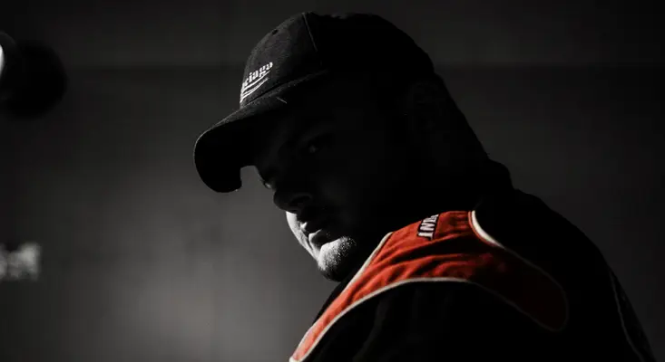 Multi-Genre Bass Producer Lizdek Unveils Studio Album ‘DUALITY’ Multi-genre bass music producer Lizdek looking over his shoulder in a dark, moody studio setting wearing a black Balenciaga cap and a red and black racing jacket.