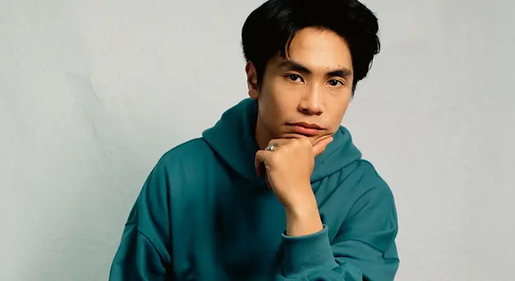 Rakhano's next chapter focuses on artistic growth and original storytelling. Close-up portrait of music artist Rakhano wearing a teal hoodie, posing with his hand under his chin against a neutral background.
