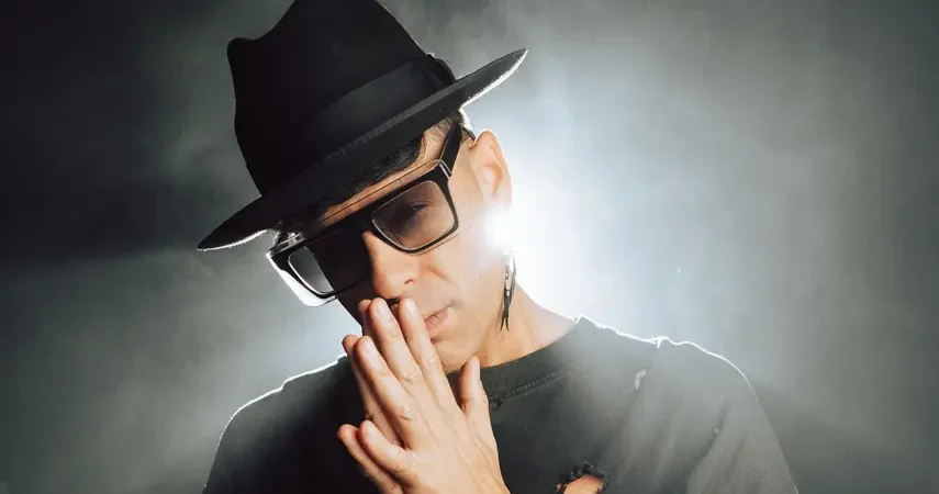 Timmy Trumpet wearing a black hat and glasses, posing with hands together in a smoky, backlit setting for his single Roses.