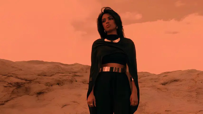 Singer Debbii Dawson wearing a futuristic black outfit with gold metallic belts standing on a desert landscape with an orange sky in the Mars music video.