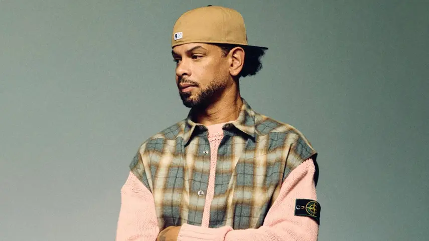 MK (Marc Kinchen) Serves Housey Vibes Through “Zone” House music producer MK (Marc Kinchen) looking to the side, wearing a tan baseball cap, a plaid vest over a pink knit sweater, posing against a solid grey background.