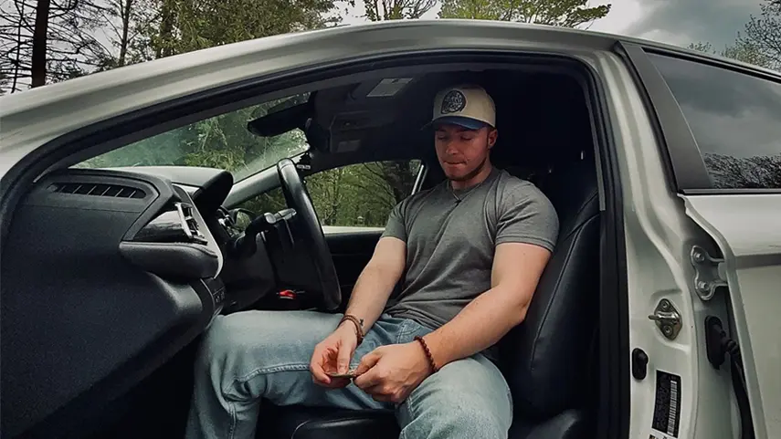 Maine independent artist KiingWavvyy, wearing a baseball cap and grey t-shirt, sitting in a white car, featured in his single “Still Want Us.”