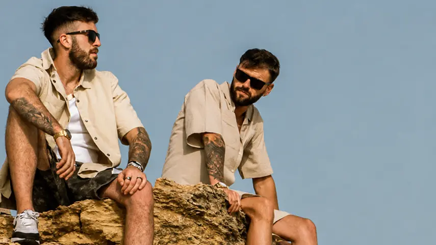 Symmetrik duo sitting on rocky cliff in beige summer outfits promoting Stay With Me summer EDM video.
