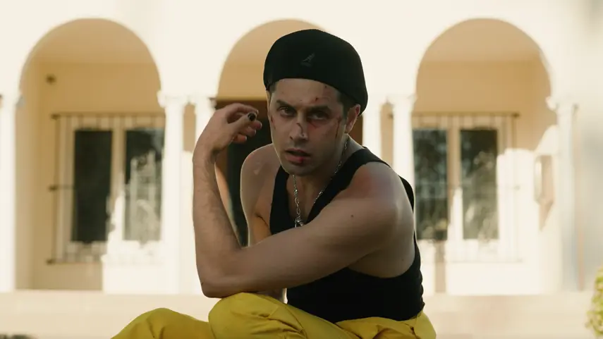 Stewart Taylor Drops "Dark Summer," A Haunting Single And Video Stewart Taylor, wearing a black tank top, black head wrap, and yellow pants, crouches with fists raised in a fighting stance, with visible cuts and bruises on his face and arm, in front of an arched Mediterranean-style building — a still from the "Dark Summer" music video.