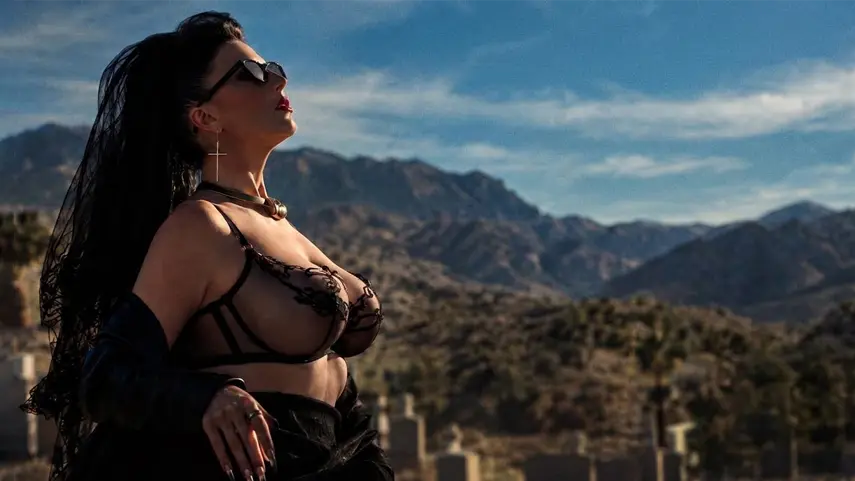 Desire poses in a black lace outfit and sunglasses in a desert cemetery scene from the "Just Another Love Song" music video.