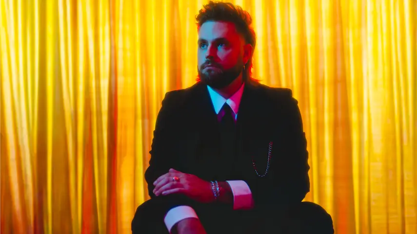 Micah Martin Reveals Debut Album's Tracklist With Single “VHS” Micah Martin wearing a black suit against a gold curtain backdrop, promotional image for his new single VHS.