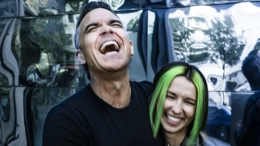 Robbie Williams and Miss Monique share a laugh while promoting their new single "Beauty In Us." Robbie Williams and Miss Monique laughing together during Beauty In Us collaboration.