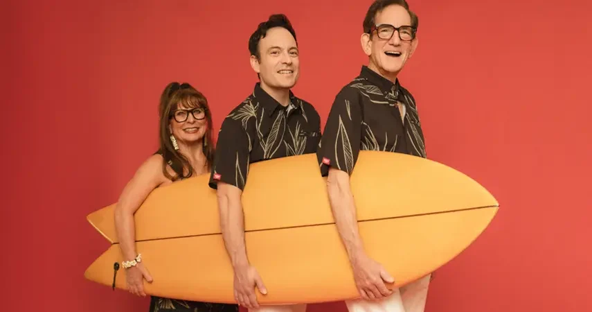 Slack Key ʻOhana family trio smiling and holding an orange surfboard against a red background.
