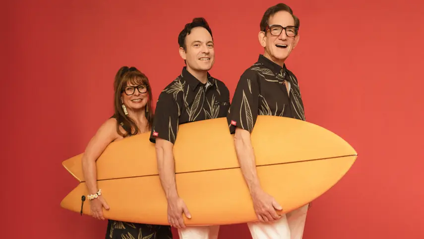Slack Key ʻOhana family trio smiling and holding an orange surfboard against a red background.