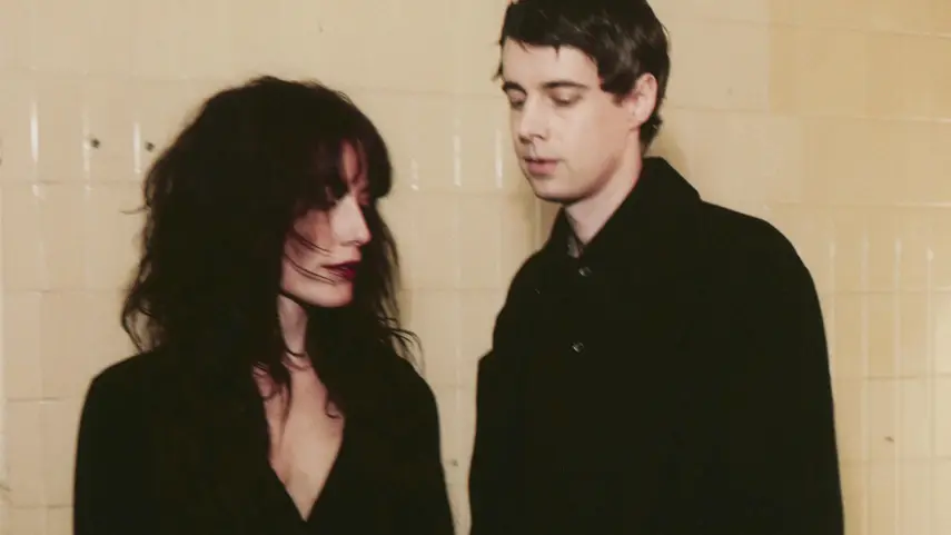 Electro-Pop duo Boy Harsher, Jae Matthews and Augustus Muller, dressed in black against a cream tiled wall.