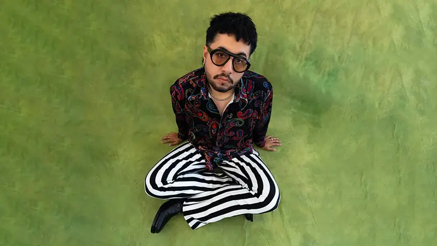 Disco funk music artist Never Dull sitting on a green backdrop in striped pants and a paisley shirt.