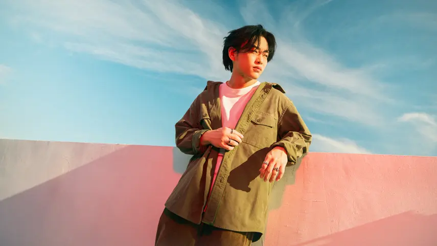 Max Jordan on the story behind his EP 'Scared to Fall.' Indie pop artist Max Jordan in an olive overshirt leaning against a pink wall under a blue sky.